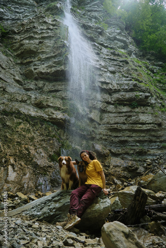 Young woman near waterfall with two dogs. Front view. Wanderlust, travel pets concept. Caucasian female traveler with australian and german shepherds enjoy nature while walking around the reserve.