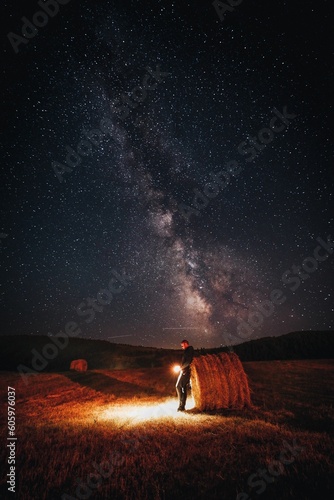 Man leaning on a hay bale roll with a light in his hands below the wonderful starry night sky