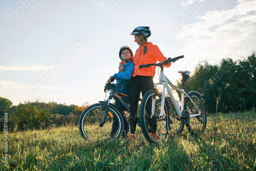 Fototapeta Naklejka Na Ścianę i Meble -  Mother and son ride bike outdoors. Happy cute boy in helmet to riding a bike in park on green meadow at sunset time. Family weekend. mothers Day