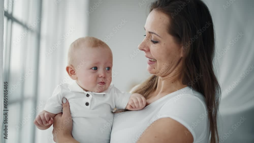 Portrait of happy family affectionate positive mom kissing baby holding in hands looking at window. Mother kisses kid at home. Motherhood, parenting, babycare, childhood, babyhood, love care concept. Portrait of happy family affectionate positive mom kissing baby holding in hands looking at window. Mother kisses kid at home. Motherhood, parenting, babycare, childhood, babyhood, love care concept.
