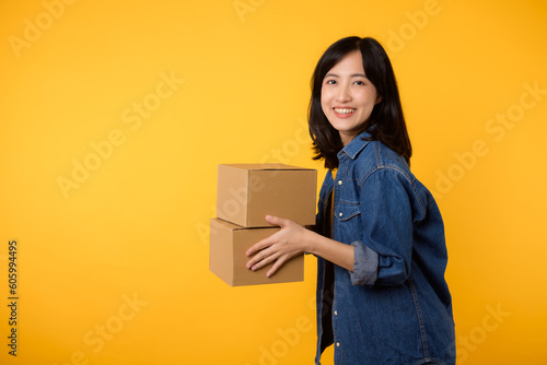 Portrait asian young woman wearing yellow t-shirt and denim shirt holding parcel box isolated on yellow studio background, Delivery courier and shipping service concept. © Jirawatfoto