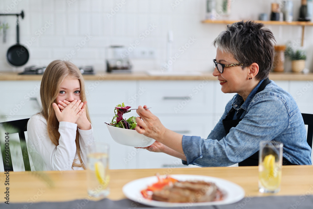 Little girl child closes her mouth and does not want to eat vegetable salad for lunch, adult mother tries to feed the child.