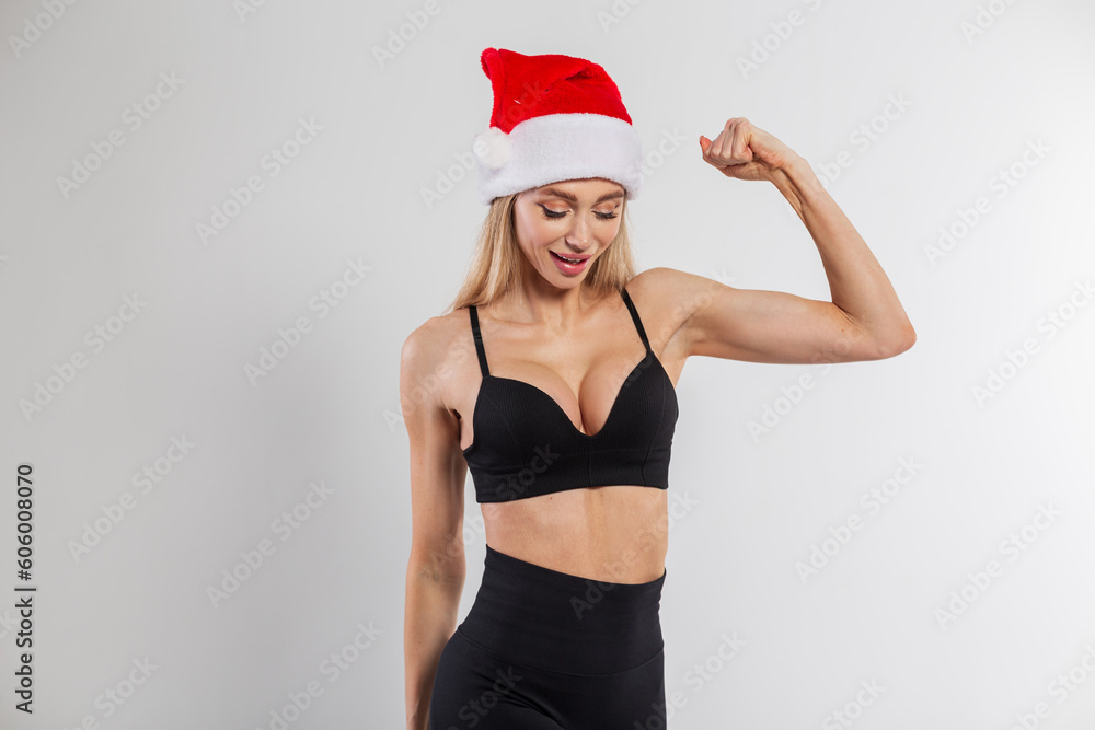 Funny beautiful fitness woman with a sexy figure in a black bra with a red santa claus hat shows biceps on a white background. Sport, beauty, health and new year Funny beautiful fitness woman with a sexy figure in a black bra with a red santa claus hat shows biceps on a white background. Sport, beauty, health and new year