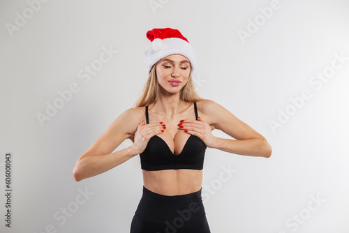 Beautiful sexy fitness lady with a slender body in a black bra with a red santa claus hat posing on a white background. New Year, sports and beauty. Girl holding her hands on breast