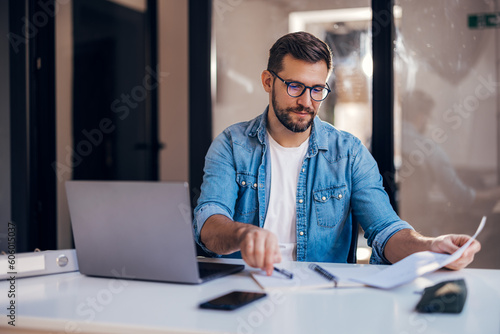 Foto Satisfied young man with glasses sitting at a desk and doing paperwork at his workplace