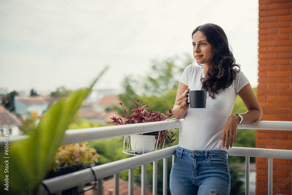 A beautiful young woman standing on the balcony holding a cup of coffee. - obrazy, fototapety, plakaty 