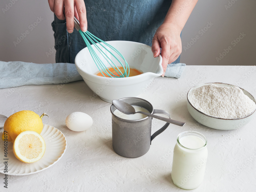 Woman beating eggs with a whisk in a bowl. Ingredients to prepare a dessert or pastry recipe. Flour, milk and lemon on the table. - obrazy, fototapety, plakaty 