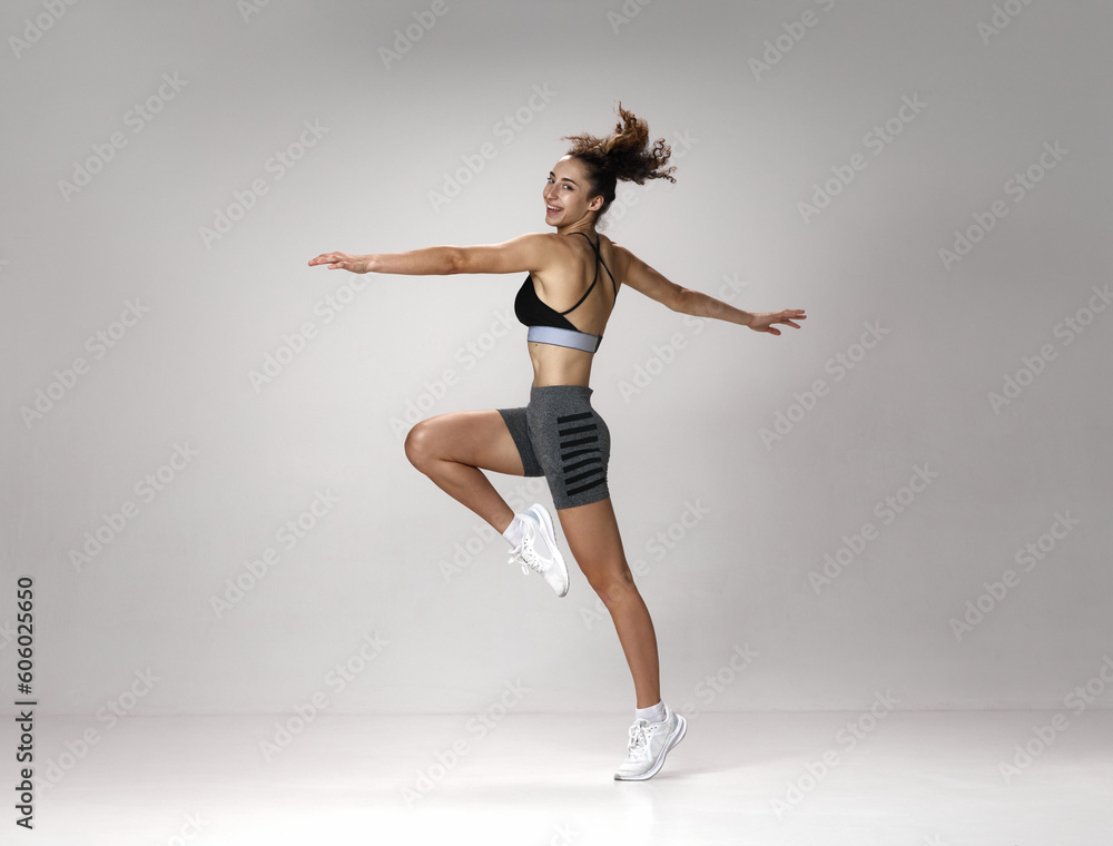 Dynamic image of happy, smiling, fit young girl in sportswear training against gray studio background. Wellness. Concept of sportive lifestyle, beauty, body care, fitness, health