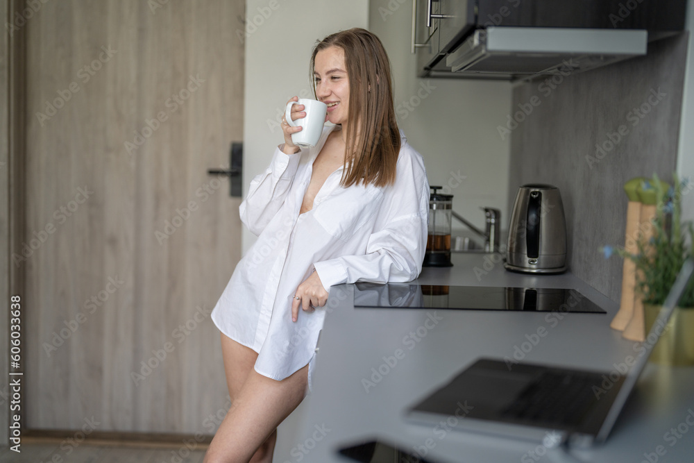 Beautiful Young Girl is Standing in the Kitchen and Drinking Coffee. Morning Routine. Wearing White shirts. Blurry Laptop and Mobile Phone on the Desk. Beautiful Young Girl is Standing in the Kitchen and Drinking Coffee. Morning Routine. Wearing White shirts. Blurry Laptop and Mobile Phone on the Desk.