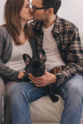 Happy couple hugging and kissing at home sitting on the couch.A French Bulldog puppy is on their lap.Family and animal life concept.
