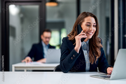 A smiling female employee talking on a mobile phone while working on a laptop at the office.