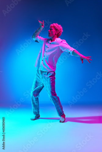 Full-length portrait of mature man in white shirt and checkered pants dancing against blue studio background in pink neon light. Concept of human emotions, lifestyle, youth, facial expression
