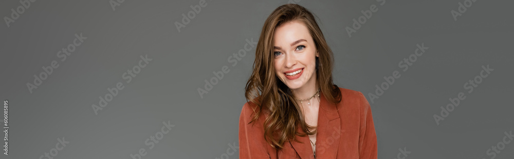 happy young woman with brunette and wavy hair posing in trendy and oversize suit with terracotta blazer and smiling while looking at camera on grey background, stylish pose, trendy chic, banner happy young woman with brunette and wavy hair posing in trendy and oversize suit with terracotta blazer and smiling while looking at camera on grey background, stylish pose, trendy chic, banner
