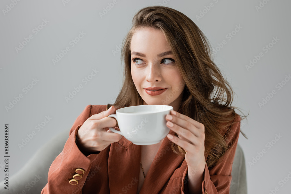 pleased young woman with long hair wearing terracotta and trendy suit with blazer and looking away while holding cup of coffee near blurred comfortable armchair on grey background in studio pleased young woman with long hair wearing terracotta and trendy suit with blazer and looking away while holding cup of coffee near blurred comfortable armchair on grey background in studio
