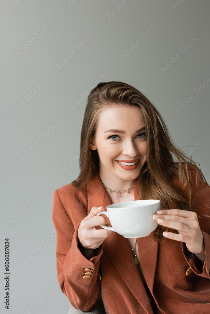 Portrait of smiling and fashionable young woman in terracotta jacket and necklaces holding cup of cappuccino and looking at camera isolated on grey Portrait of smiling and fashionable young woman in terracotta jacket and necklaces holding cup of cappuccino and looking at camera isolated on grey