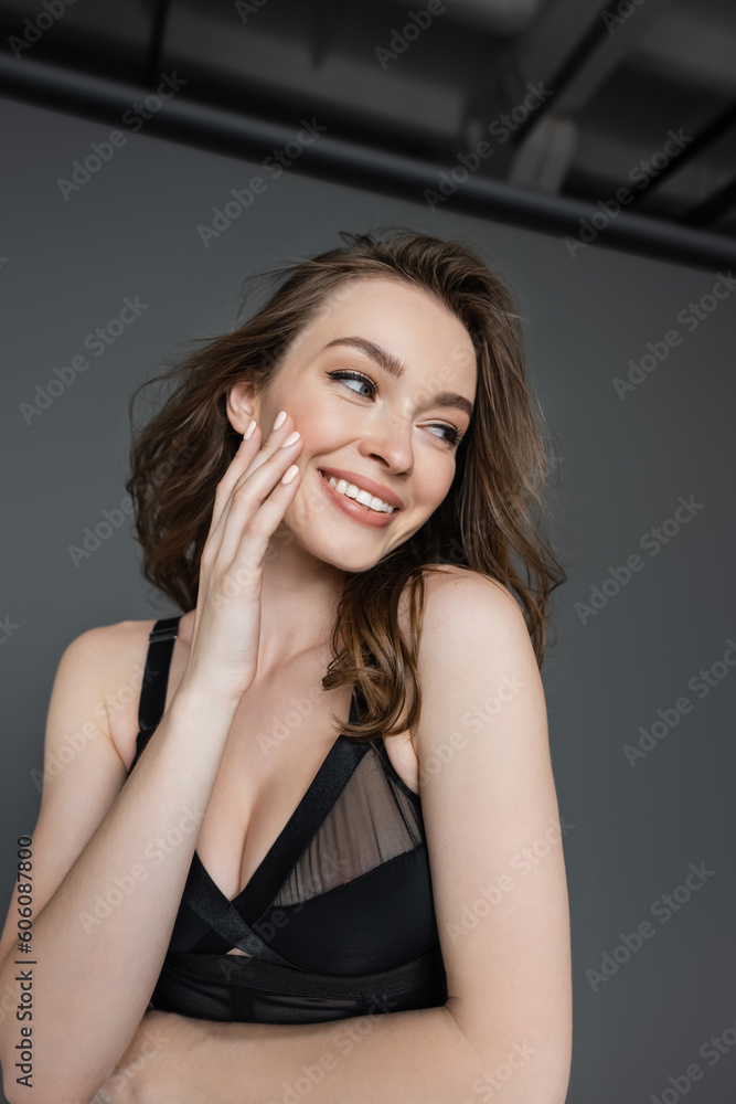 Portrait of fashionable young brunette woman with natural makeup wearing sexy black bodysuit while touching face and looking away and standing on grey background
