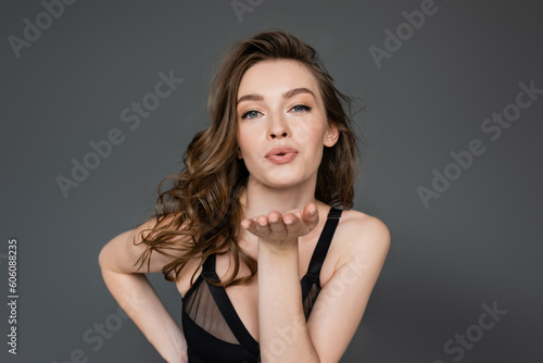 Seductive young brunette model with natural makeup and hairstyle wearing black bodysuit and looking at camera while blowing air kiss and flirting isolated on grey