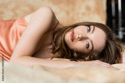 graceful young woman with brunette hair lying in silk slip dress and looking at camera on blurred and linen fabric on mottled beige background, sophistication, sensuality, elegance
