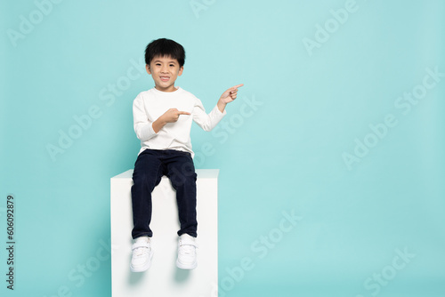 Asian little boy sitting on white box and pointing to empty copy space isolated on green background, Full body composition