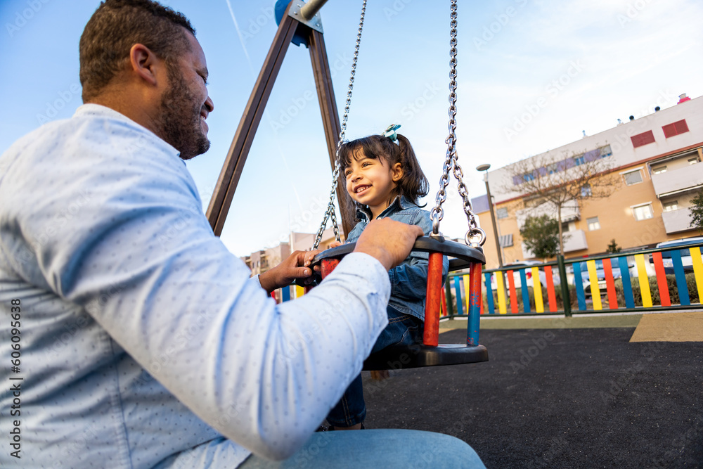 Obraz premium A dark-skinned father happily plays with his daughter on an outdoor playground. The father pushes the daughter who is on a children's swing from the front. Concept of love in single-parent families.
