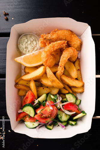 Lunch tempura shrimp fried breaded with lemon, rustic potato wedges, vegetable salad and sauces in a paper box.
