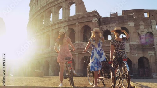 Three happy young women friends tourists with bikes waving hats at Colosseum in Rome, Italy at sunrise.