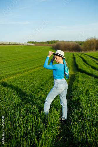A cute woman in jeans and a hat stands in a green field. A smiling woman in a blue top and jeans walks in the green grass. © Дмитрий Ткачук