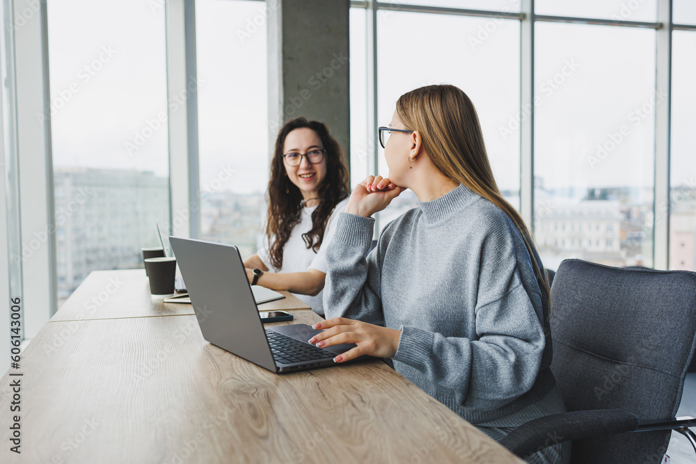 Beautiful two young confident business women sitting in an office room working on a laptop computer. The work of colleagues in a modern office. Beautiful two young confident business women sitting in an office room working on a laptop computer. The work of colleagues in a modern office.