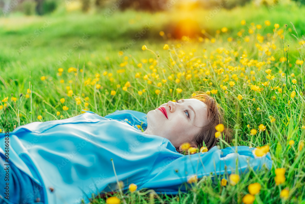 Side view happy young woman in blue blouse lays on a green grass lawn with yellow flowers, looks up and smiles. Female enjoy the moment, relaxing outdoors at sunset. Spring or summer season concept. Side view happy young woman in blue blouse lays on a green grass lawn with yellow flowers, looks up and smiles. Female enjoy the moment, relaxing outdoors at sunset. Spring or summer season concept.