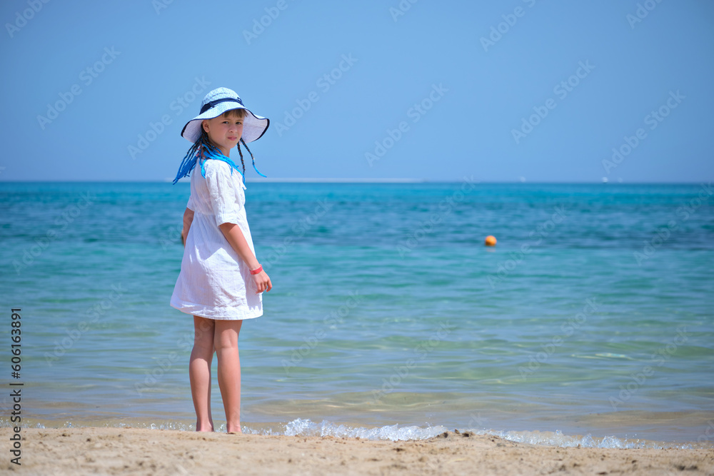 Rear view of child girl in white summer dress and sunshade hat with blue ribbons in long braids standing barefoot on sandy beach enjoying sea view
