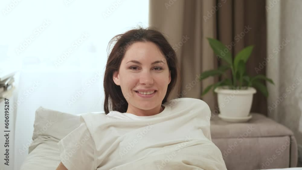 A lazy pretty young woman is sitting on a bed in the early sunny morning, smiling and looking at the camera. A lazy pretty young woman is sitting on a bed in the early sunny morning, smiling and looking at the camera.