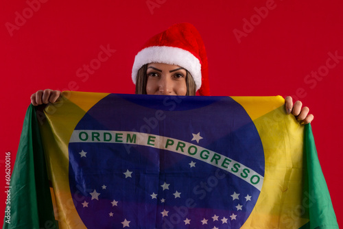 Very beautiful adult woman with the flag of Brazil, wearing a Santa Claus hat and dress, in a studio photo with a red background, with space for text and making various facial expressions photo