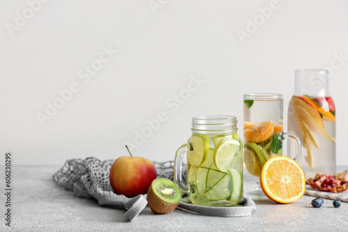 Glass, mason jar and bottle of infused water with different sliced fruits on grey table