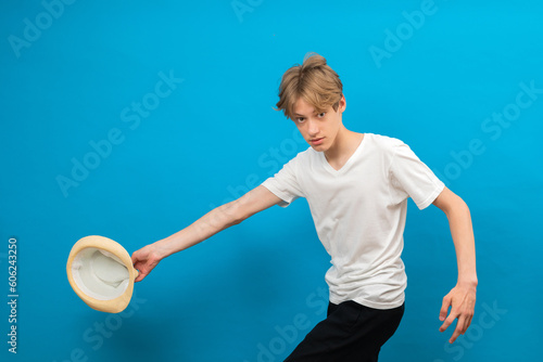 Cheerful energetic teenager boy running and holding white hat in hand in studio on blue background © nazariykarkhut