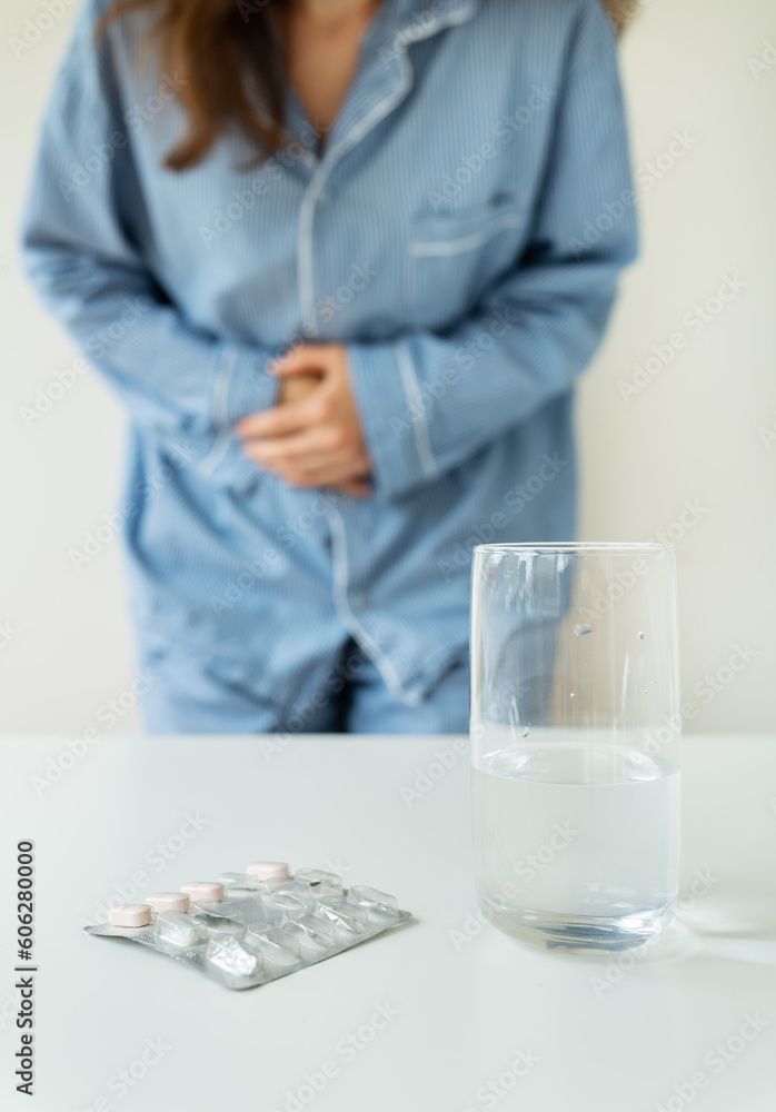 A young girl in pajamas stands and holds on to her stomach, there is a glass of water and pills on the table. The concept of PMS, indigestion, migraine, stress, depression. A young girl in pajamas stands and holds on to her stomach, there is a glass of water and pills on the table. The concept of PMS, indigestion, migraine, stress, depression.