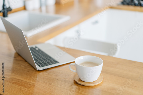 Cup of cofee and laptop on wooden table, view on white kitchen in modern style