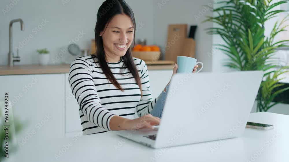 Video of smiling young woman doing video call with laptop while holding a cup of coffee in the kitchen at home. Video of smiling young woman doing video call with laptop while holding a cup of coffee in the kitchen at home.