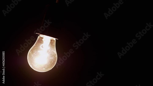 Hanging Swinging Glowing Light Bulb in a Dark Room on the Black Background. Old Edison tungsten incandescent bright shining on and tunning off. Energy crisis. Electricity. Power outage. Blackout. photo