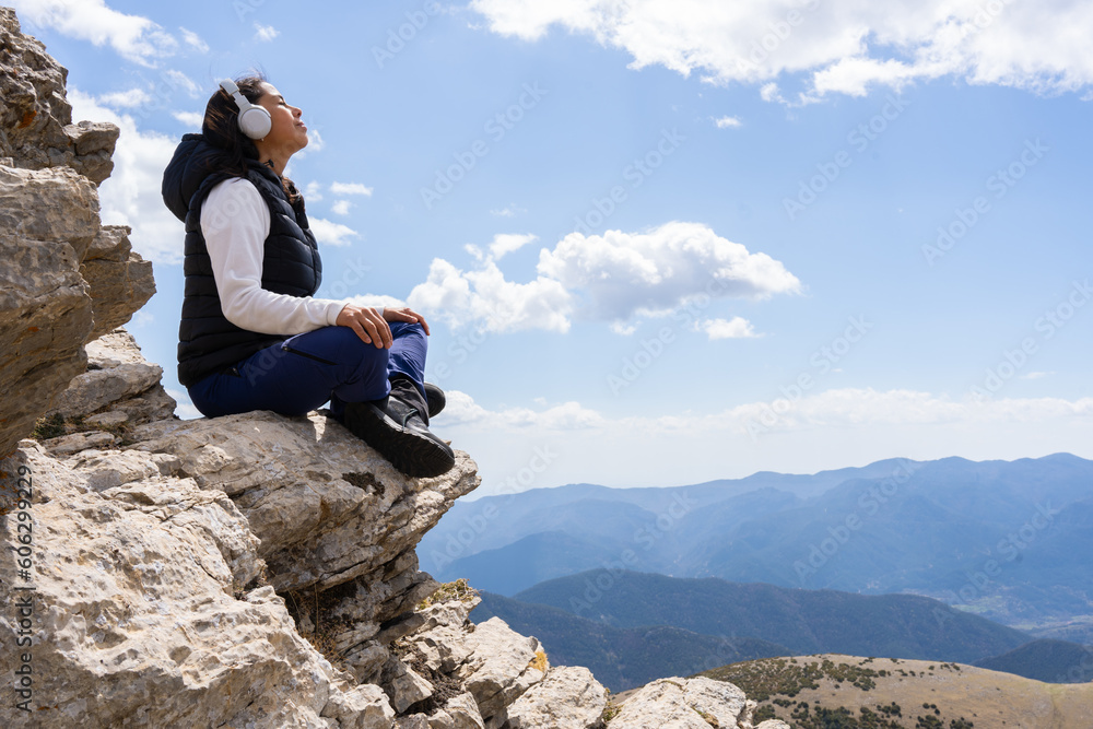 Latin woman relaxes listening to music with her headphones sitting on a rock on top of the mountain after a long day of trekking and hiking