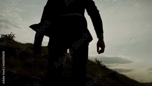 Rear view of an adult man in a business suit fluttering in the wind climbs a steep and rocky hillside with long steps against the backdrop of the rays of the sunset. Concept of achieving the goal