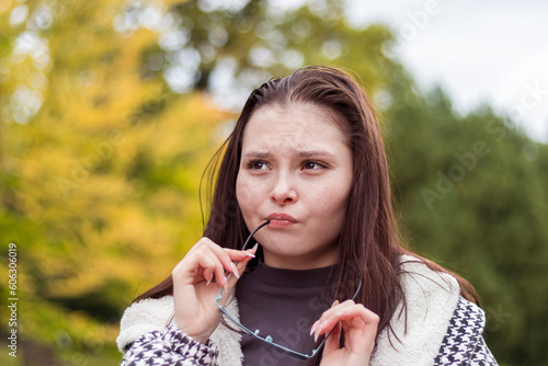 A young woman stands in a city park, thoughtfully touching her chin with glasses, thinking about an important choice.