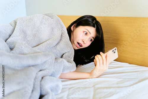 寝室で朝の寝起きの起床時にスマートフォンのアラームに驚き寝坊して出勤に遅刻しそうな1人暮らしの女性 photo