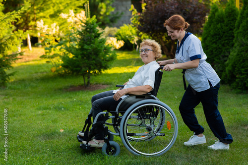 Caucasian female doctor walks with an elderly patient in a wheelchair in the park. Nurse accompanies an old woman on a walk outdoors. 