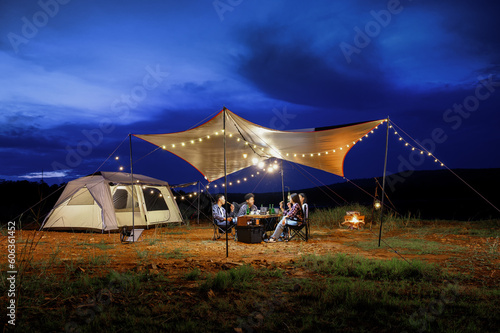 Outdoor camping tent in the forest park, party dinner with friends under tarp or flysheet and warm lighting at night near natural under a blue twilight sky. Overview of travel camping with friends.