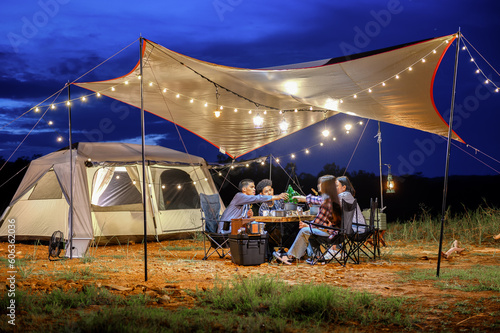 Outdoor camping tent in the forest park, party dinner with friends under tarp or flysheet and warm lighting at night near natural under a blue twilight sky. Overview of travel camping with friends.