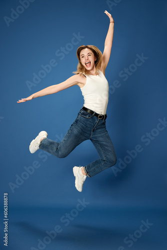 Full-length portrait of young woman in casual clothes posing, jumping in happiness and excitement against blue studio background. Concept of youth, human emotions, facial expression, success