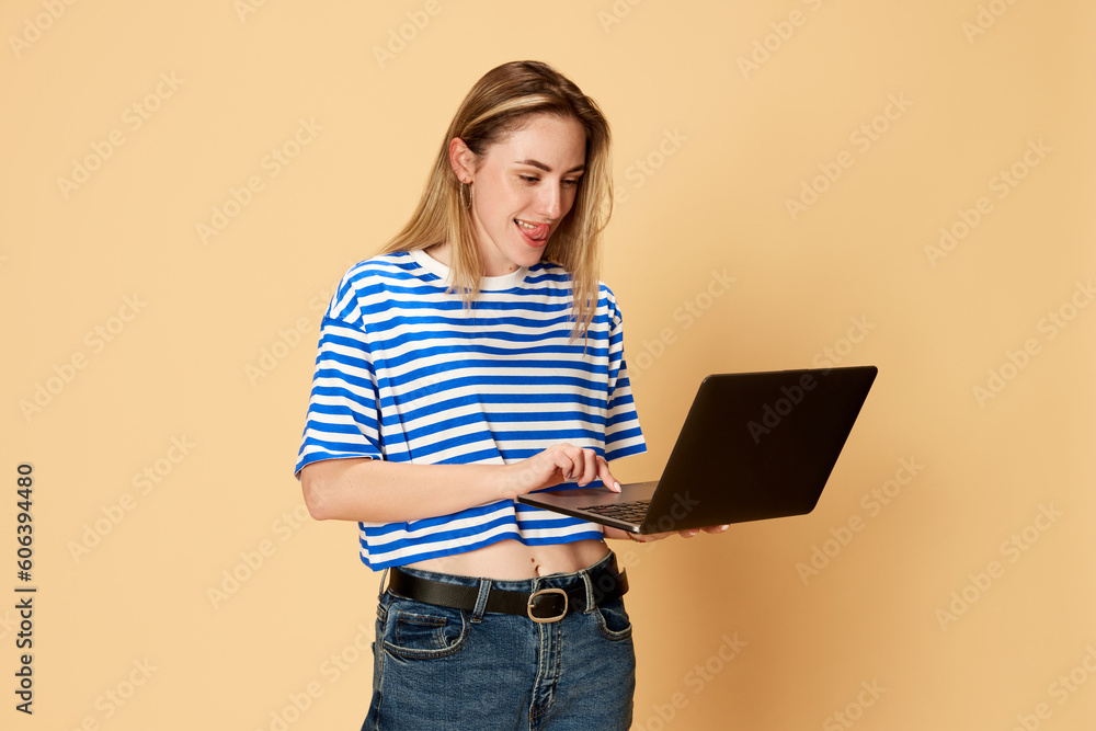 Portrait of young girl in striped shirt styanding with laptop against yellow studio background. Good job, creating projects. Concept of youth, human emotions, facial expression, business and education Portrait of young girl in striped shirt styanding with laptop against yellow studio background. Good job, creating projects. Concept of youth, human emotions, facial expression, business and education