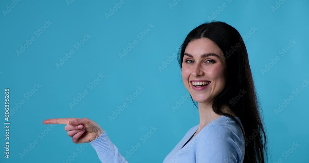 Woman standing in profile against a blue background. She turns towards the camera, shows teeth while smiling widely, and points finger forward, as if indicating something interesting ahead. Woman standing in profile against a blue background. She turns towards the camera, shows teeth while smiling widely, and points finger forward, as if indicating something interesting ahead.
