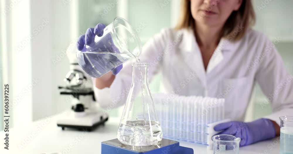Female laboratory technician in rubber gloves pours clean water into glass flask. Professional scientist mixes solutions using laboratory shaker Female laboratory technician in rubber gloves pours clean water into glass flask. Professional scientist mixes solutions using laboratory shaker