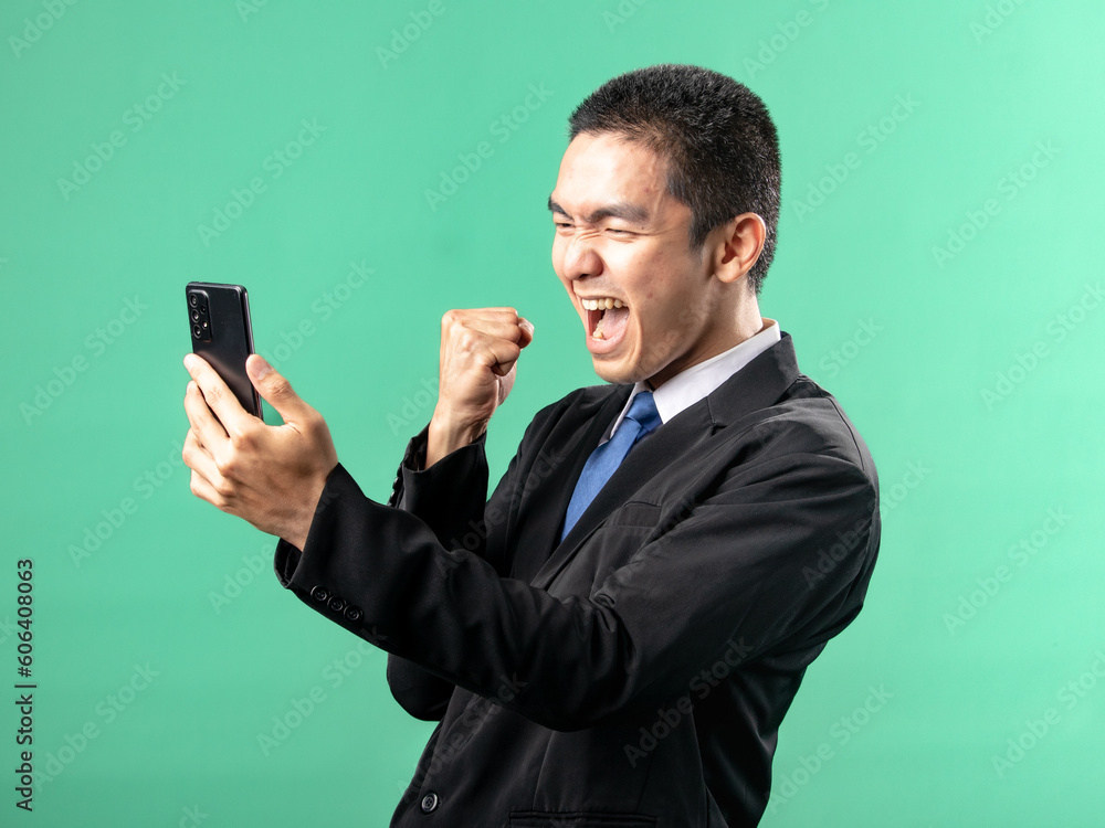 A portrait of an Asian man wearing a white shirt and a black suit, looking very happy while looking at a cellphone, isolated on a green background. A portrait of an Asian man wearing a white shirt and a black suit, looking very happy while looking at a cellphone, isolated on a green background.